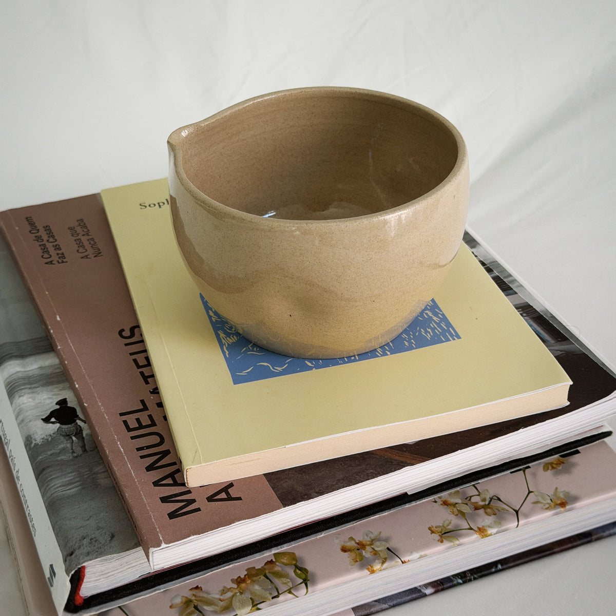 A handmade ceramic bowl with a small pouring spout, in a light tan glaze, displayed on top of a stack of books against a white fabric background.