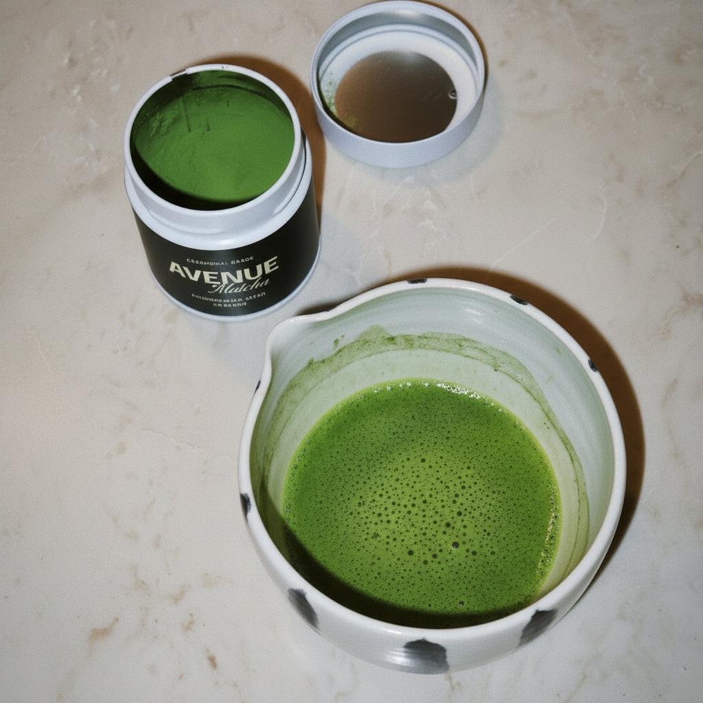 A bowl of brewed green matcha tea with a small metal strainer and an open jar of green powder on a beige marble surface. The jar label reads 'AVENUE'.