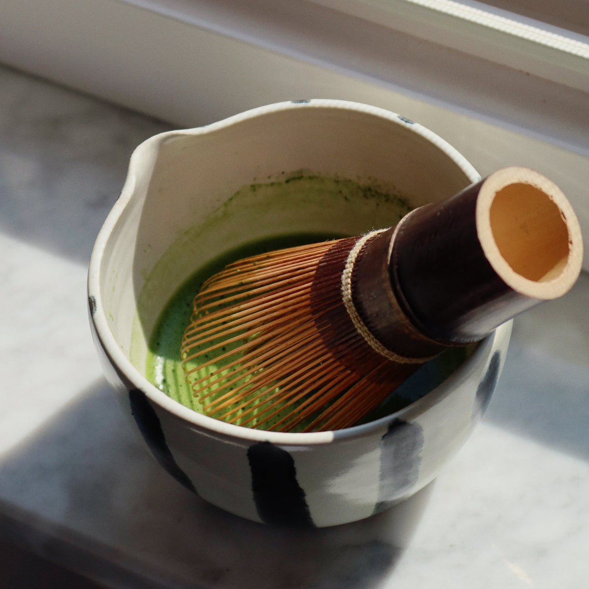 A ceramic matcha whisk bowl with black and white stripes, filled with green matcha powder and a bamboo whisk, resting on a marble surface near a window.
