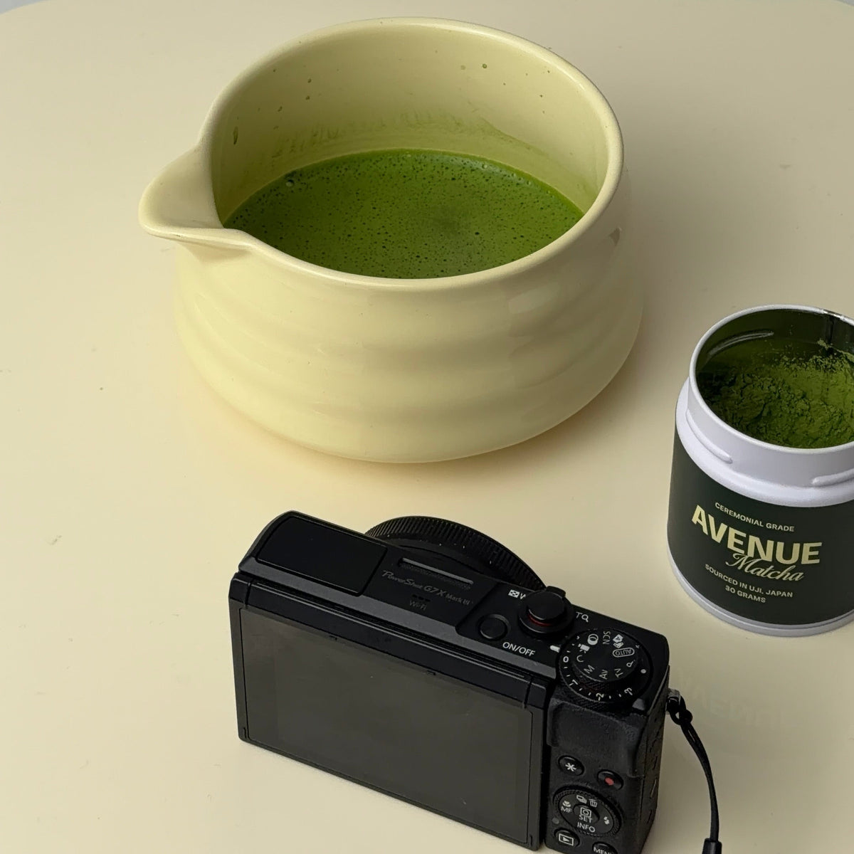 A pale yellow ceramic pitcher filled with green matcha powder sits on a cream tabletop, alongside a black compact camera and a small jar labeled 'AVENUE Matcha'.