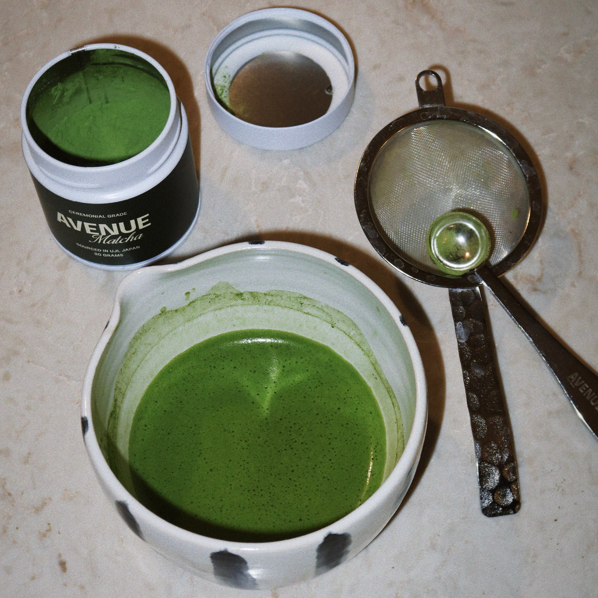 A bowl of brewed green matcha tea with a small metal strainer, an open jar of green powder labeled AVENUE, and an empty lid, arranged on a beige marble surface.