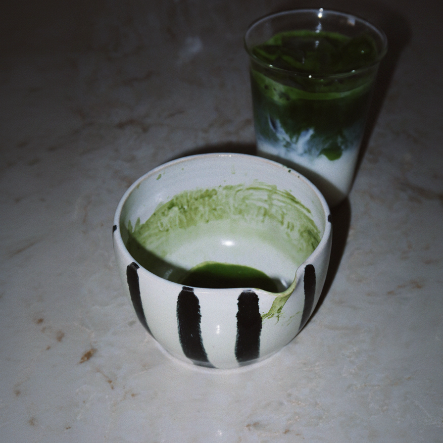 A white ceramic matcha tea bowl with black painted stripes, containing green matcha powder, with a glass of layered green and white matcha drink behind it on a light marble surface.