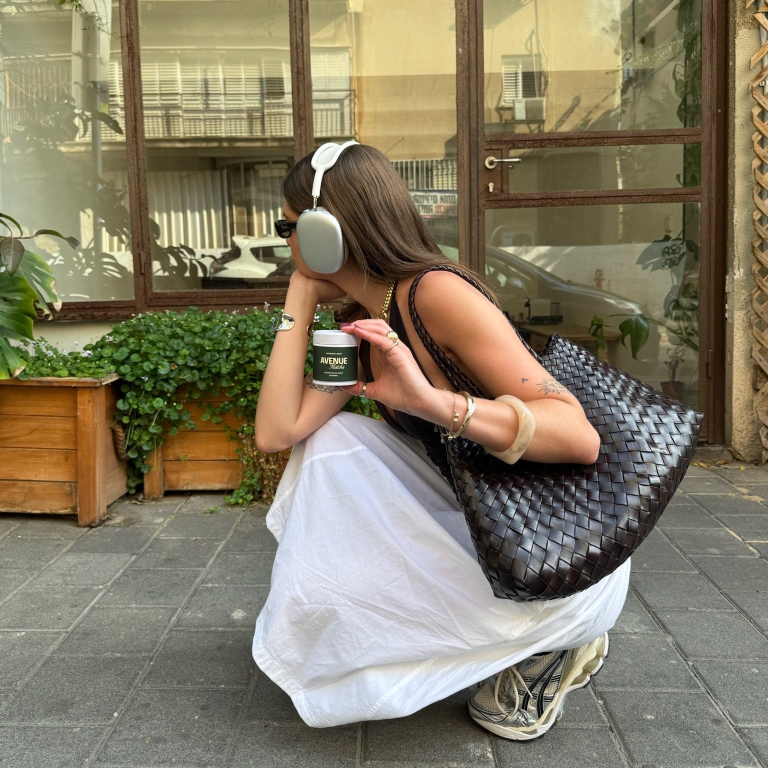Person sitting on a stone pavement holding a avenue matcha tin and a large woven bag, with a building and plants in the background.